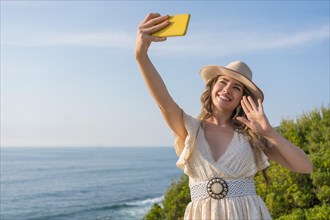 Horizontal photo of a blonde beauty woman in summer clothes waving while taking a selfie next to the sea