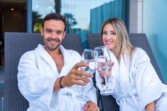 Couple celebrating a perfect honeymoon in a hotel spa toasting with white wine while looking at camera dressing white bathrobe sitting on a lounge
