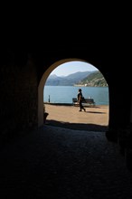 Woman Walking in an Old Beautiful Street Tunnel with Arch from Brusino Arsizio on the Waterfront in a Sunny Summer Day and with Lake Lugano and Mountain View over Morcote