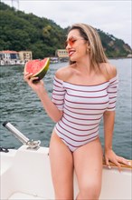Vertical photo of a blonde beauty woman eating watermelon sitting on the edge of a boat