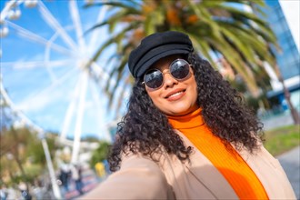 Beauty latin woman with sunglasses and hat taking a selfie in the city next to a ferris wheel in a sunny day of winter