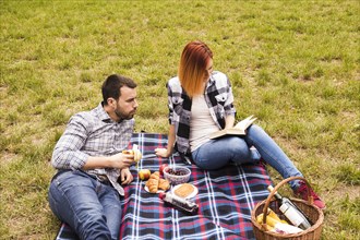 Man eating banana looking her girlfriend reading book picnic