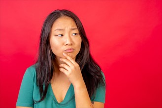 Studio photo with red background of a curious and doubtful chinese woman with hand on chin