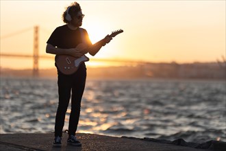 A man stands at the waterfront and playing guitar at bright orange sunset. Mid shot