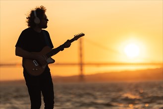 Romantic man wearing wireless headphones stands at the waterfront and playing guitar at bright orange sunset. Mid shot