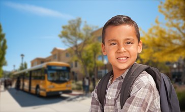 Cute young hispanic boy wearing a backpack near a school bus on campus