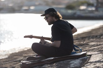 A man hipster playing guitar at the quay. Mid shot