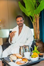 Vertical portrait of a handsome caucasian man dressing bathrobe enjoying luxury breakfast in the hotel room