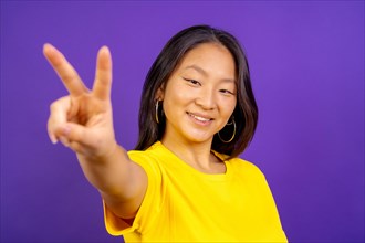 Studio photo with purple background of a smiling chinese woman gesturing peace sign with fingers