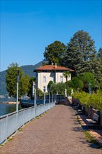 Walkway with Lamps and Railing and a House on the Waterfront on Lake Lugano with Mountain and Trees in a Sunny Summer Day in Porto Ceresio