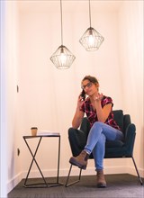 Vertical photo of a woman talking to the mobile sitting with a coffee at home in a cozy room