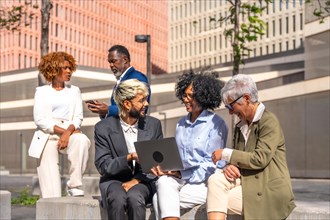 Multi-ethnic business people using laptop sitting outdoors in a bench next to a financial building
