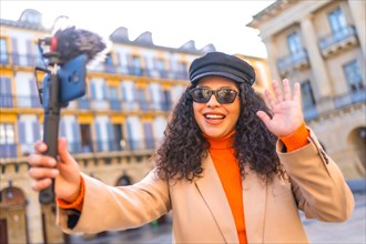 Latin stylish woman waving at camera while recording a video visiting a city in autumn