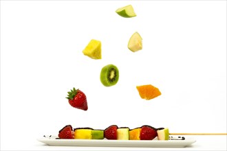 Skewer of assorted fruits covered with chocolate with pieces of fruit falling isolated on a white background