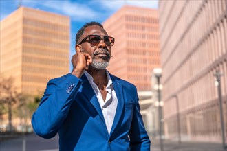 Mature african businessman with sunglasses adjusting the earphone to have a mobile conversation outdoors