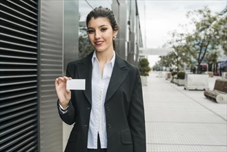 Happy young businesswoman standing outside office building showing her business card