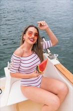 Vertical photo of a cute stylish woman sitting on a boat eating watermelon
