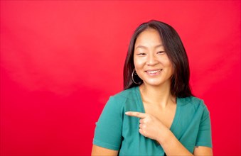 Studio photo with red background of a happy chinese woman pointing aside in a blank space