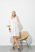 Beautiful woman in white sundress standing by wicker chair in sunny room