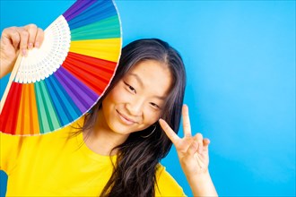 Studio photo with blue background of a chinese woman using a rainbow colored folding fan gesturing peace