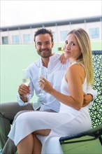 Vertical portrait of lovers celebrating love with champagne sitting on a chair in the terrace of an hotel