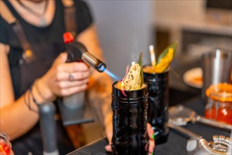 Cropped photo with close-up of a female bartender flaming the garnish of a cocktail with a torch in the counter of a bar