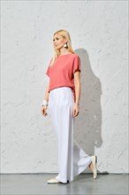 Trendy woman with blond hair in pink shirt and loose white trousers walking along white wall