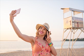 Horizontal photo of a blonde adult woman gesturing victory while taking a selfie on the beach during sunset