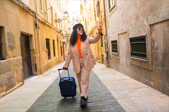 Chic latin woman wearing warm autumn clothes taking a selfie while carrying luggage along a street