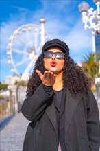 Vertical portrait of a latin stylish woman blowing a kiss standing in the city in a sunny day of winter