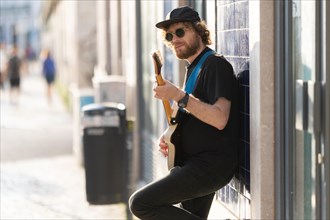 A man hipster wearing sunglasses standing by the wall and playing guitar. Mid shot