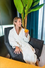 Vertical photo of a chic woman in bathrobe drinking orange juice sitting in a luxury hotel room