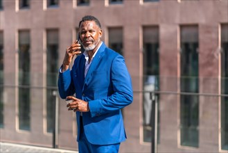 African mature businessman talking to the mobile phone outdoors while drinking a take away coffee