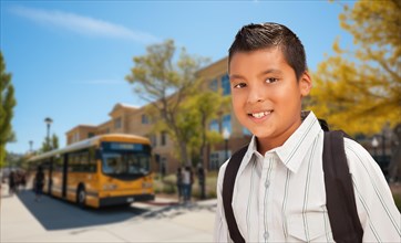 Happy young hispanic boy wearing a backpack near a school bus on campus