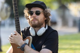 A man hipster wearing sunglasses and wireless headphones sitting in the park with his guitar