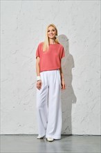 Cute woman with blond hair in pink shirt and loose white trousers standing by textured wall