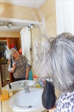 Mature white-haired woman drying her hair with a hand-held hair dryer in front of a mirror