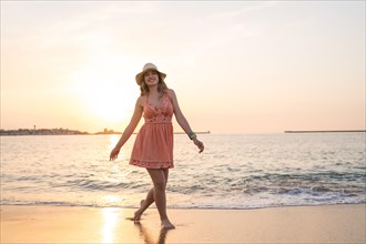 Happy woman in summer clothes walking along the beach at the edge of the water during sunset