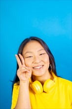 Vertical studio photo with blue background of a chinese woman with headphones gesturing victory sign