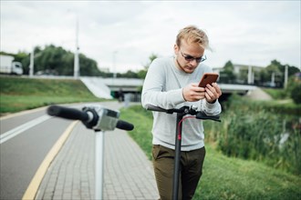 Young handsome blond looking at the route on his smartphone to go with a friend on a trip on electric scooters