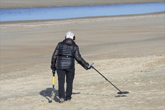 Elderly woman with metal detector beachcombing on sandy beach along the North Sea coast in winter