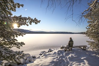 Tourist sitting on bench looking over snow-covered frozen lake in winter