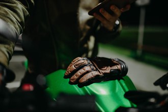 A brown leather motorcyclist glove lies on the tank of a green motorcycle while the motorcyclist looks at the travel route