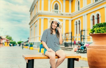 The woman is smiling wearing a hat while sitting on a bench calling on the phone with the background out of focus