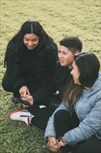 Three individuals of Hispanic-Latino ethnicity sitting on the lush green grass of a park