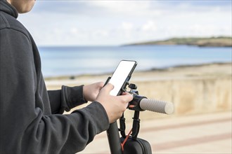 Person holding a smartphone while standing with a scooter by the sea