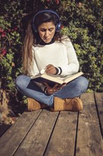 Woman with headphones focused on reading a book while sitting on a wooden deck amidst greenery with sunlight