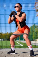 Beautiful young fitness girl warms up with a rubber expander before training at the stadium. Attractive slender brunette in an orange tracksuit. Active lifestyle