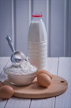 Close up view eggs flour in bowl scooping bottle of milk on white varnished old wooden board eating and drinking concept