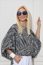A cheerfully smiling woman in a loose black-and-white patterned blouse and sunglasses leaned her hand against the wall and looked away